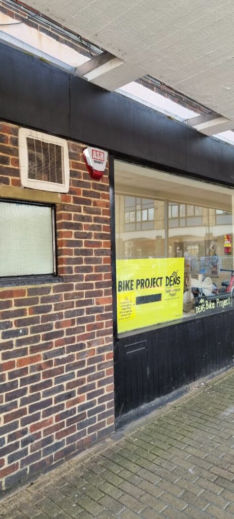 ASH Security alarm system on the exterior of the DENS Bike Project shop, featuring a prominent yellow sign displaying "BIKE PROJECT DENS."