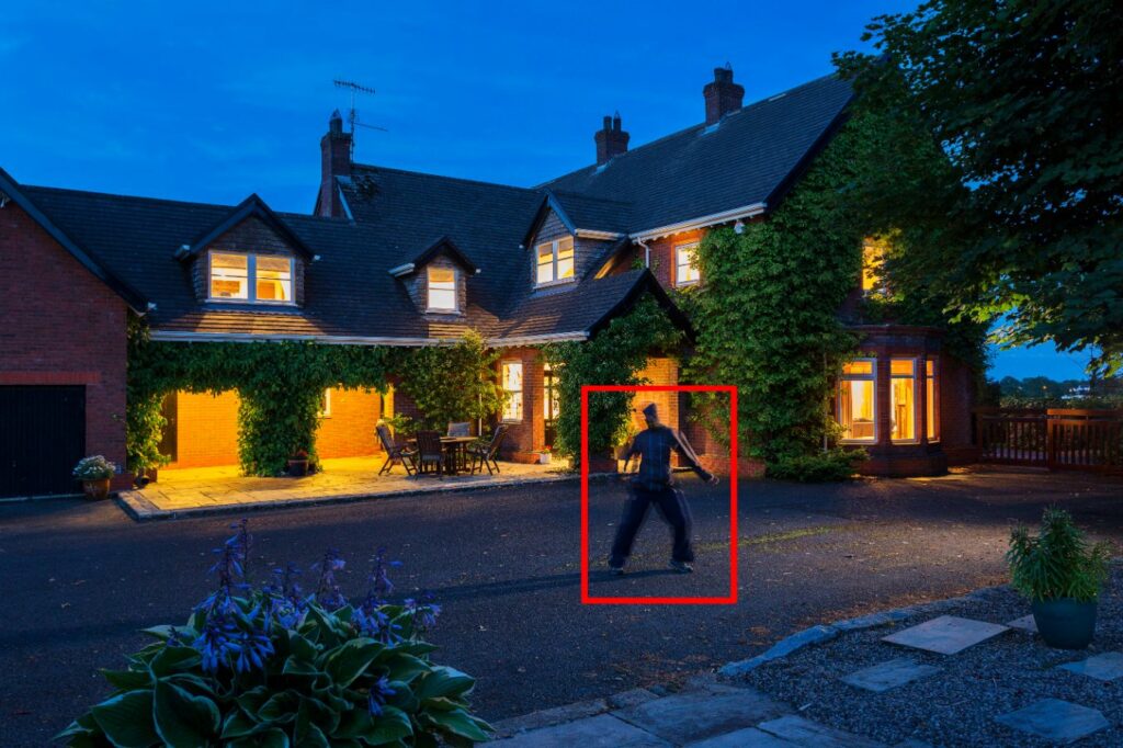 Intruder in dark clothing walking near a well-lit residential property, highlighting the importance of monitored CCTV for home security.