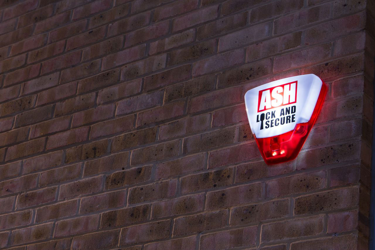 ASH Security illuminated alarm box mounted on a brick wall, featuring the text "ASH LOCK AND SECURE," highlighting security solutions for businesses and homes.