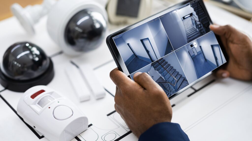 Person using a tablet to monitor security camera footage, with CCTV cameras and a motion sensor in the foreground, illustrating modern security solutions for homes and businesses.